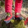 Mann steht mit beiden Beinen mit roten Forstschuhen auf mosbewachsenem Baum, daneben eine Axt am Baum