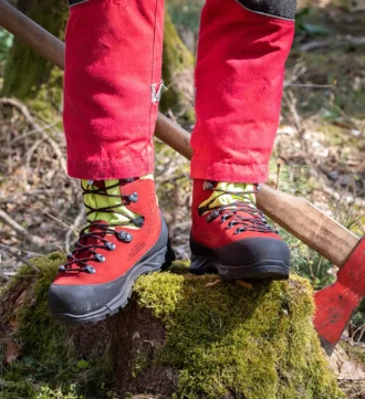 Mann steht mit beiden Beinen mit roten Forstschuhen auf mosbewachsenem Baum, daneben eine Axt am Baum
