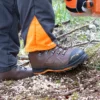 Mann steht mit beiden Beinen mit brauen Forstschuhen auf auf der Erde, während er mit der Motorsäge arbeitet.