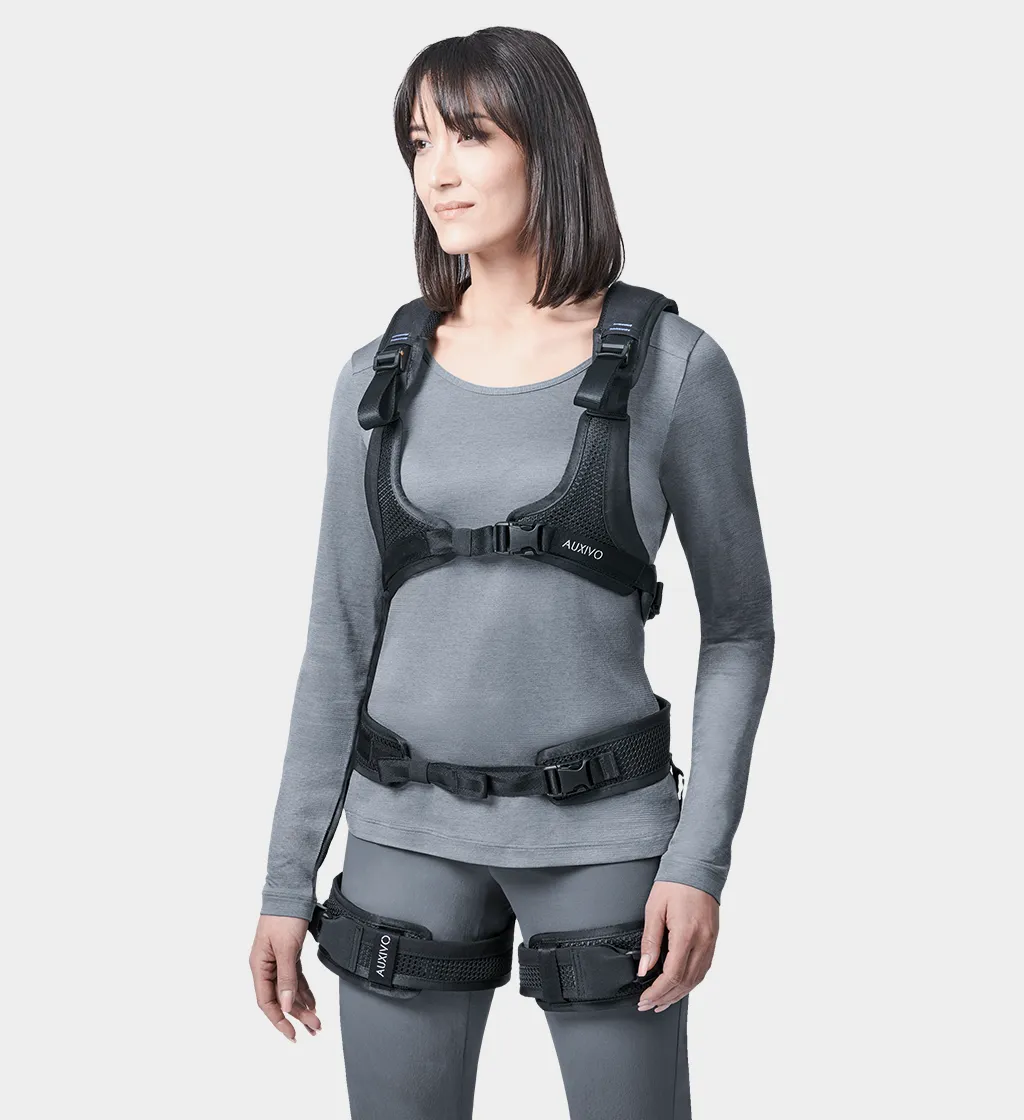 Frontale Ansicht einer Frau in grauem Oberteil und grauer Hose, die ein schwarzes Auxivo Exoskelett trägt, das ihren Rücken und ihre Hüften unterstützt