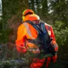 Rückansicht einer Person in leuchtend oranger Forstschutzkleidung und Helm, die im Wald an einem Baum arbeitet und dabei ein schwarzes Auxivo LiftSuit Exoskelett trägt.