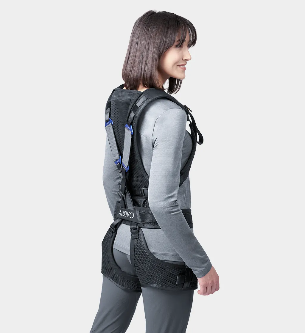Seitliche Ansicht einer Frau in grauem Oberteil, die ein schwarzes Auxivo Exoskelett trägt, das ihren Rücken und ihre Hüften unterstützt.