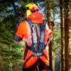 Rückansicht einer Person in oranger Forstschutzkleidung und Helm, die in einem Wald arbeitet und dabei ein schwarzes Auxivo OmniSuit Exoskelett trägt
