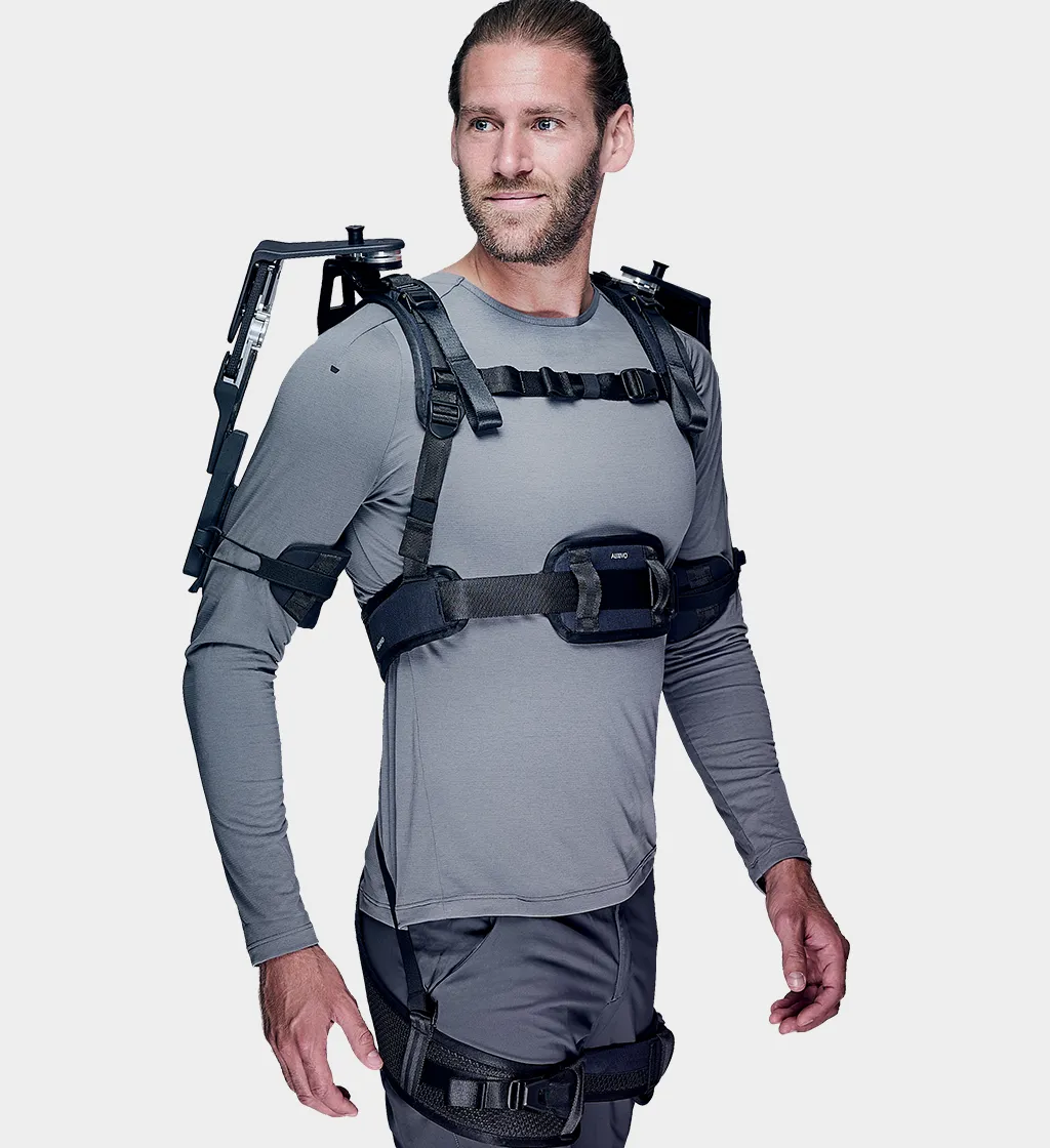 Dreiviertelansicht eines Mannes in grauem Oberteil und grauer Hose, der ein schwarzes Auxivo OmniSuit Exoskelett trägt, das seine Schultern, seinen Rücken und seine Hüften unterstützt.